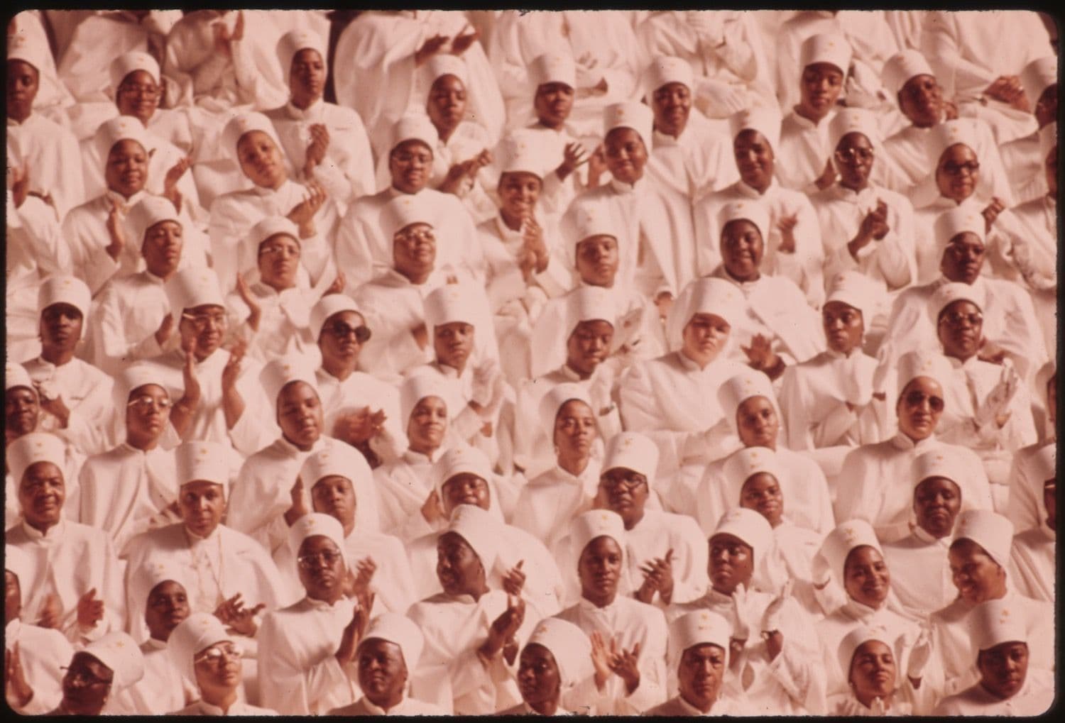 A large group of African American people in white robes and head coverings, likely a choir, densely packed together and raising their hands.