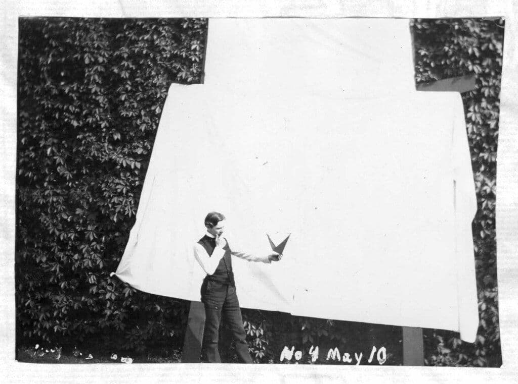 A vintage black and white photograph of a man interacting with two birds in flight, set against a backdrop of trees and a bright sky.