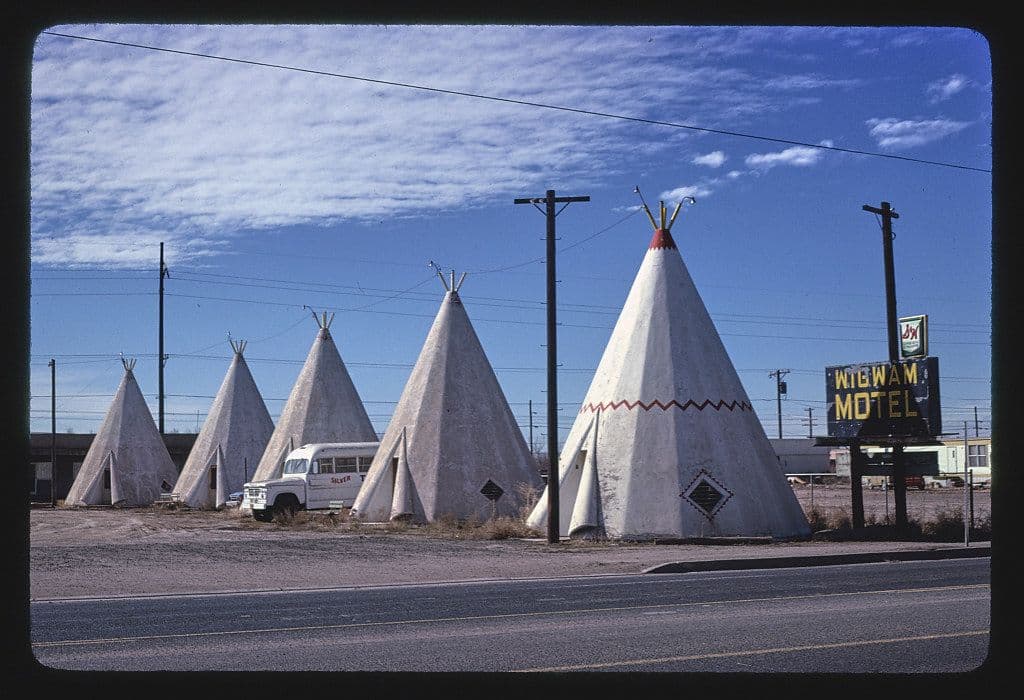 A row of concrete wigwams that make up the Wigwam Motel. The wigwams are painted a light gray color and have decorative patterns painted on them. A white van is parked in front of the wigwams. A sign that reads "Wigwam Motel" is visible. The sky is blue with some clouds. A road runs in front of the wigwams.